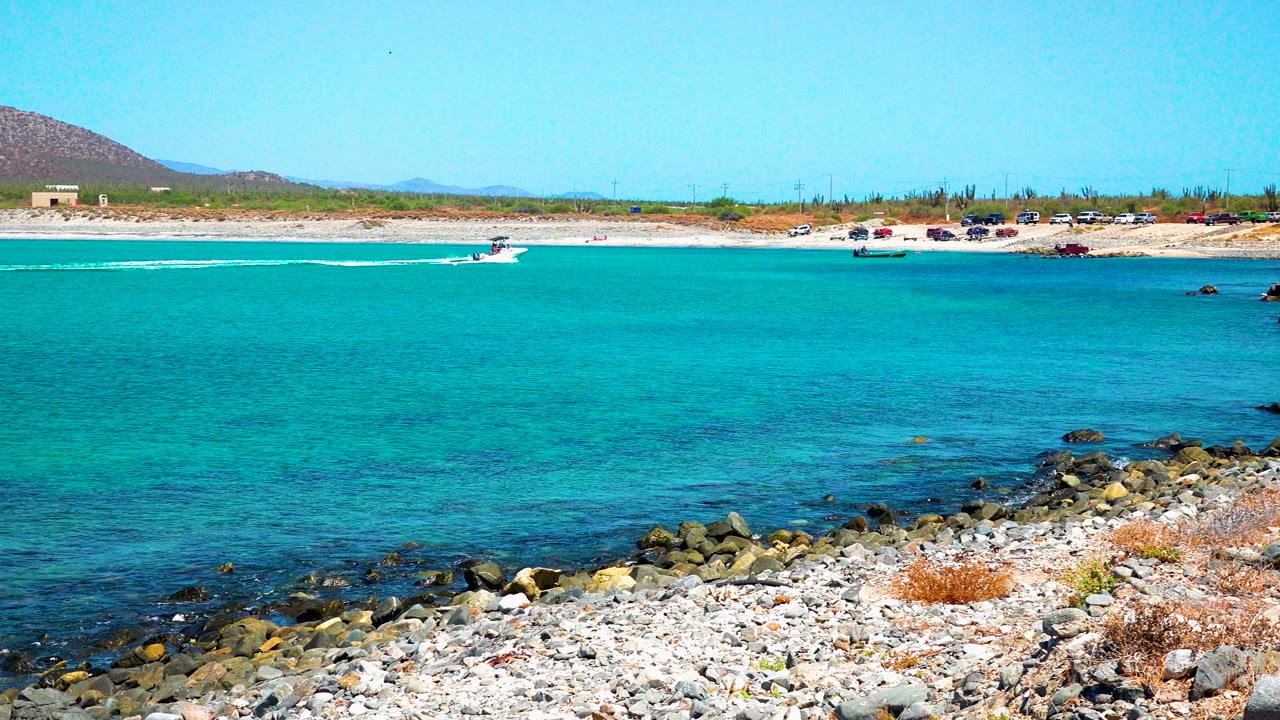Rocky foreground and clear turquoise water at El Sargento La Ventana, Mexico, with a small boat near the sandy far shore and no visible people.