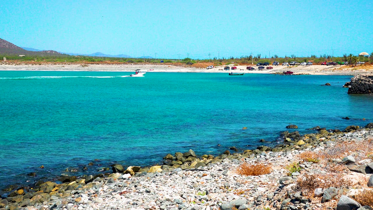 A small speedboat moves across the turquoise water at El Sargento La Ventana, Mexico, with rocks in the foreground, beach in the distance, and no visible people.