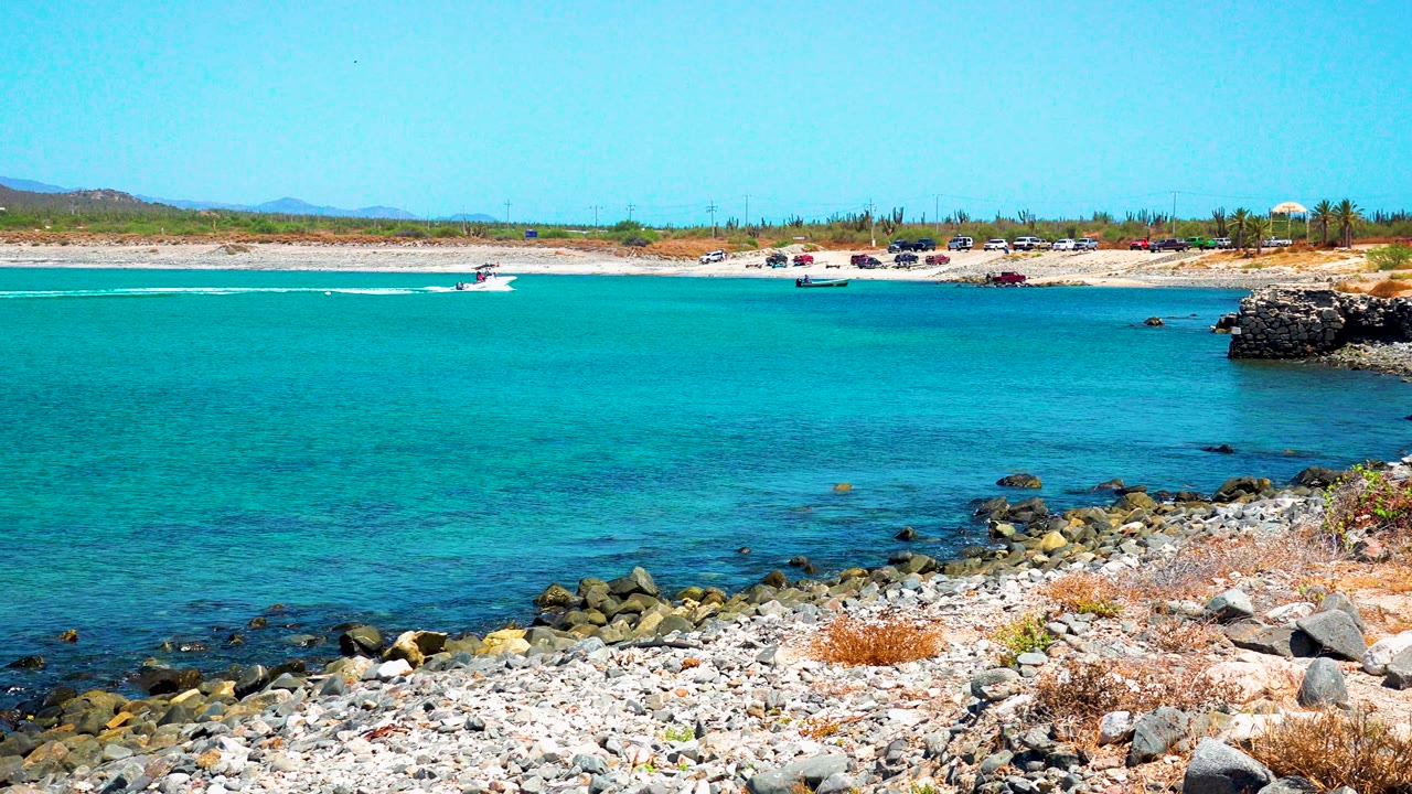 The El Sargento La Ventana shoreline appears with pebbles, shoreline rocks, bright turquoise water, a distant boat wake, and no visible people.