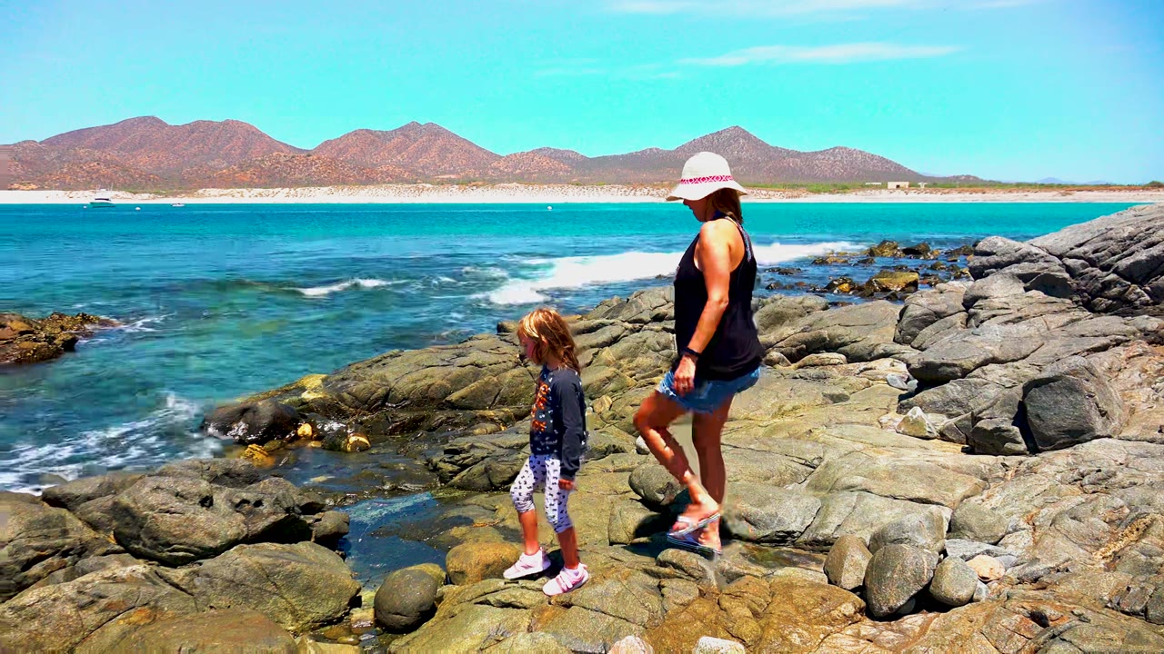 At 0:00 in El Sargento, Mexico, a woman in a white hat and a child step across sunlit rocks with blue ocean, surf, and mountains behind them.