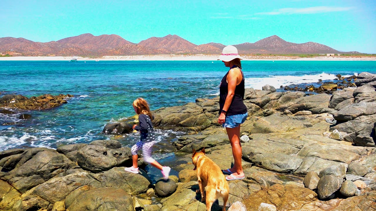 At 0:02 in El Sargento, Mexico, the child steps ahead across the uneven rocks while the woman stands to the right and the tan dog moves through the foreground.