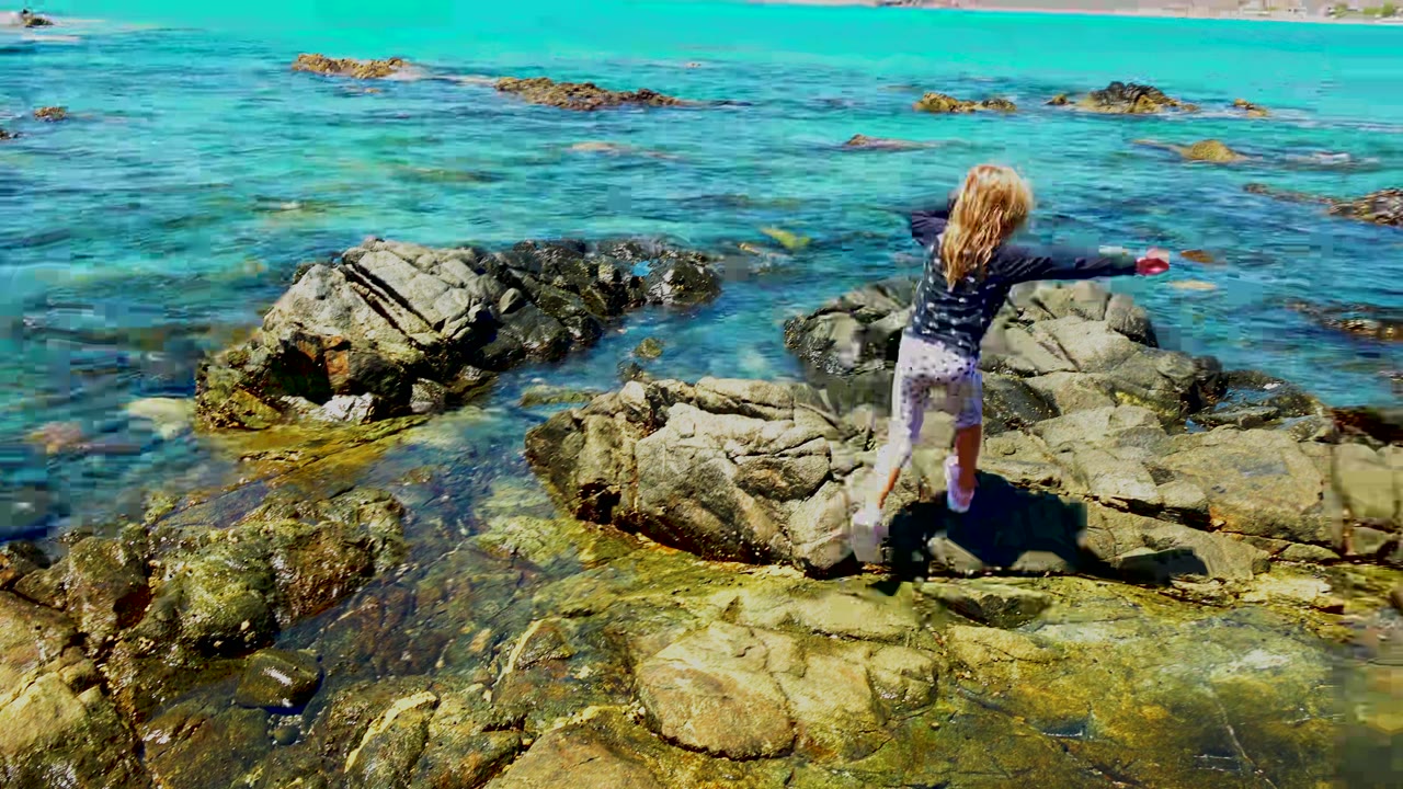 A young girl steps carefully across uneven shoreline rocks in El Sargento, Mexico, with shallow turquoise water and distant mountains behind her.