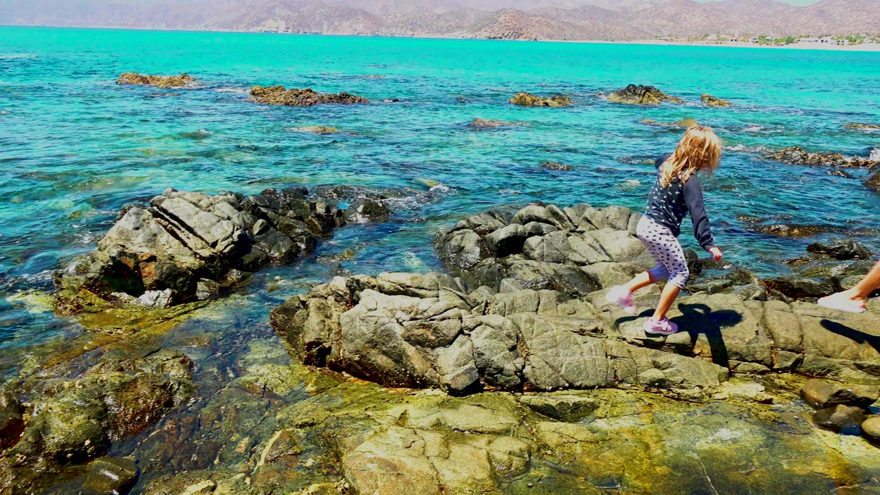 Girl balancing on shoreline rocks near clear blue water.