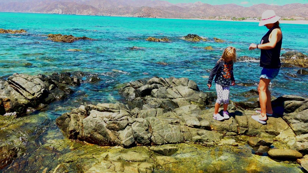 A woman in a wide sun hat appears at the right as the girl stands on the rocks in El Sargento, Mexico, with clear water and low mountains in the background.