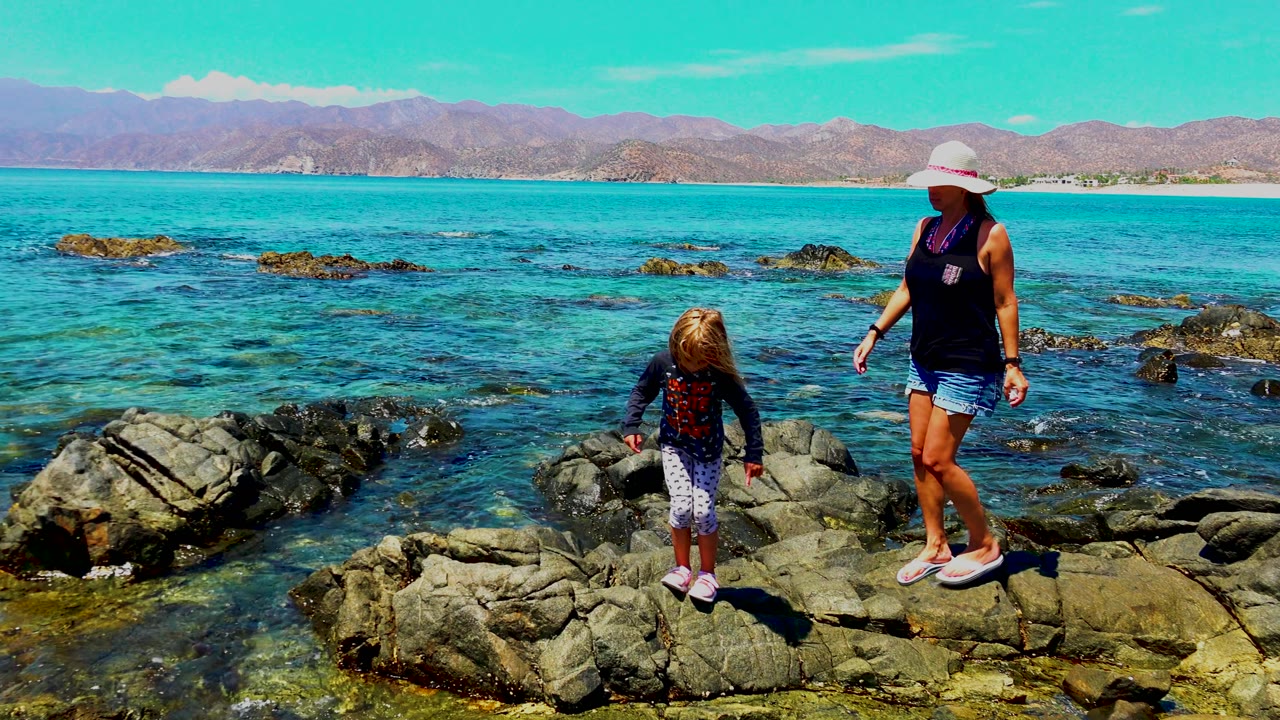 A woman in a white sun hat and a child stand on dark rocks at the La Ventana shoreline, with clear blue ocean and distant mountains behind them.