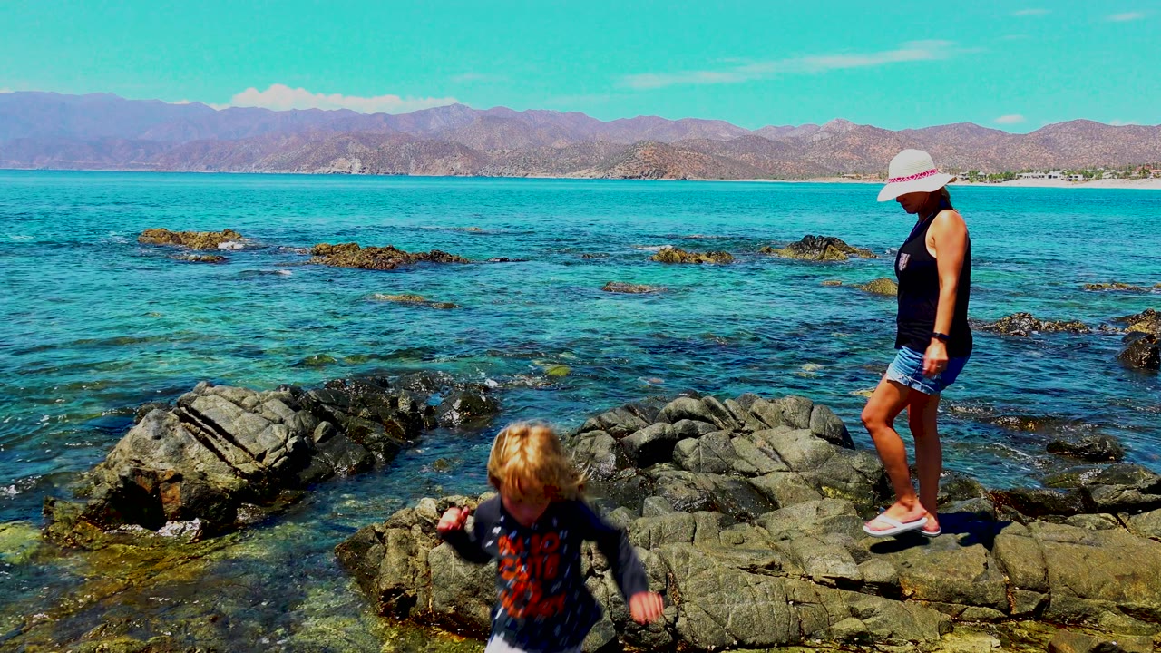 The child moves toward the lower right edge of the rocks while the woman stands above on the La Ventana shoreline, backed by blue water and mountain ridges.