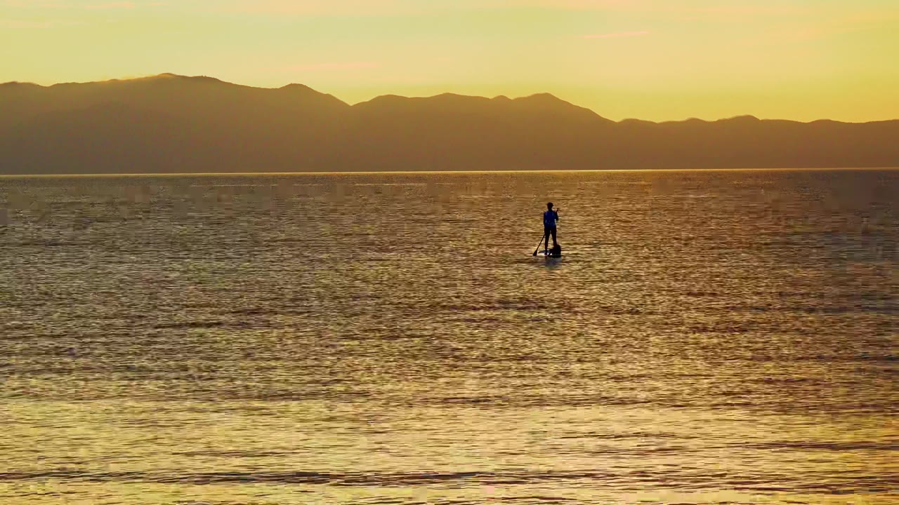 A distant silhouetted paddleboarder stands near the center of golden ocean water in El Sargento La Ventana, Mexico, with dark mountains along the sunset horizon.