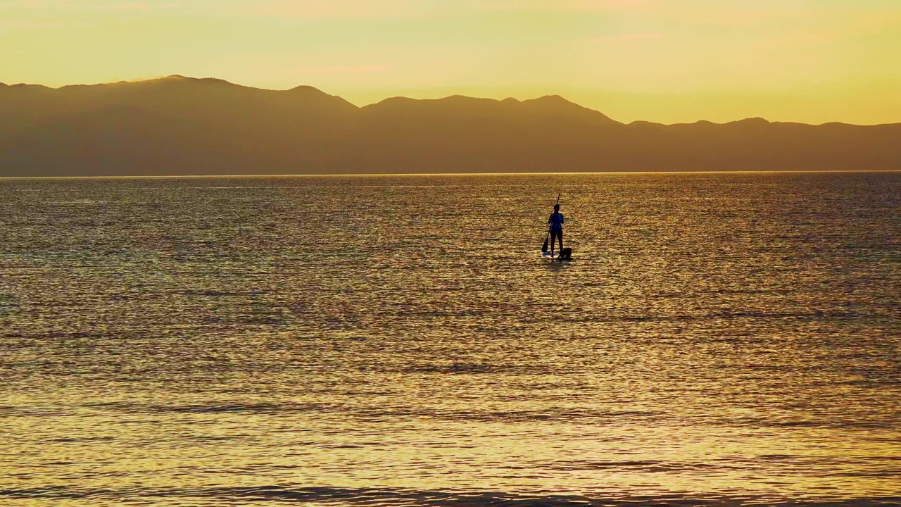 Paddleboarder silhouetted on calm sea under a yellow sunset sky.