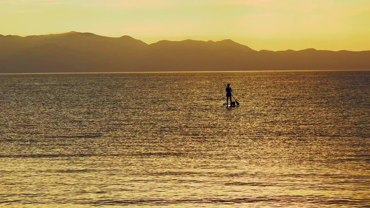 A small paddleboarder silhouette sits slightly right of center on bright reflected ocean water in El Sargento La Ventana, Mexico, with layered mountains stretching across the background.