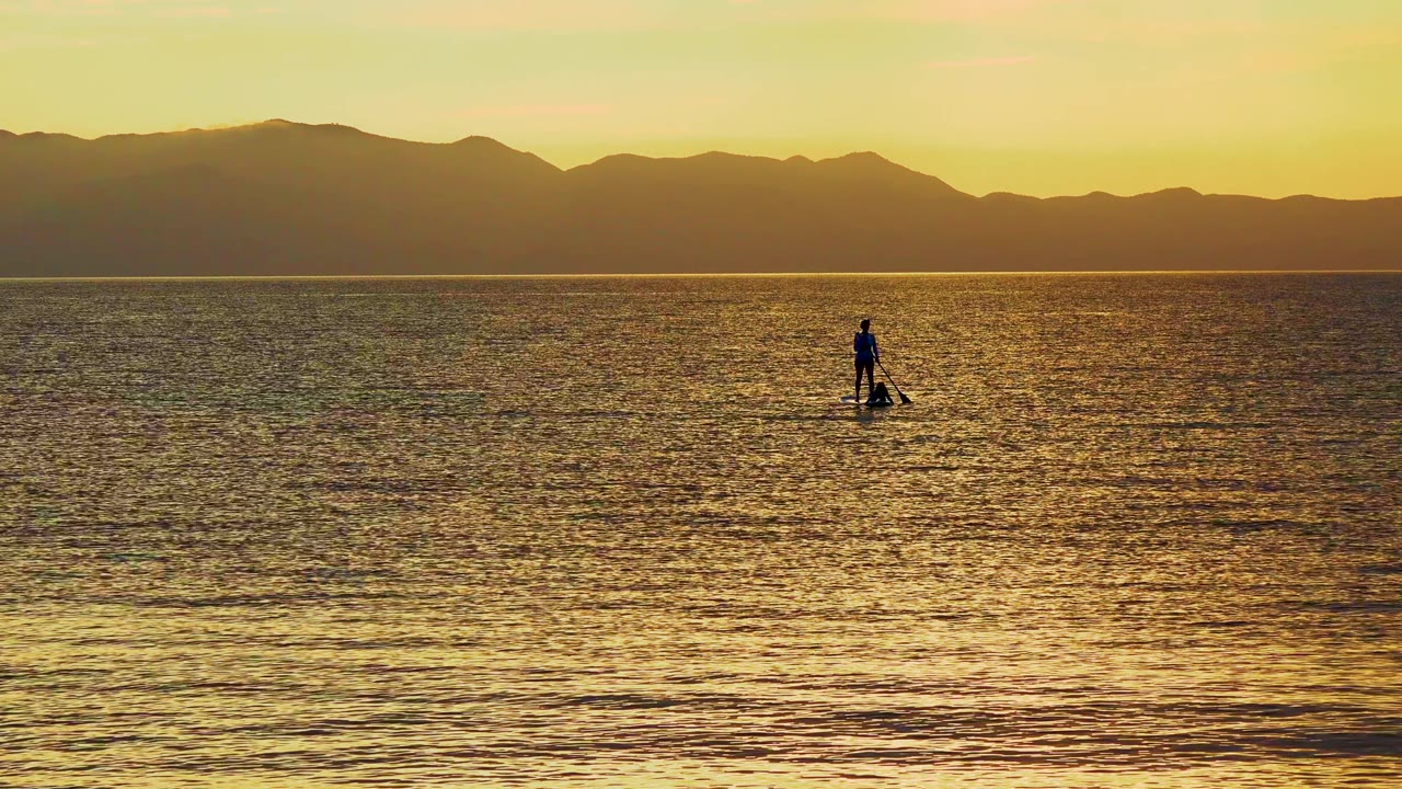 Paddleboarder on reflective ocean water below a dark mountain skyline.