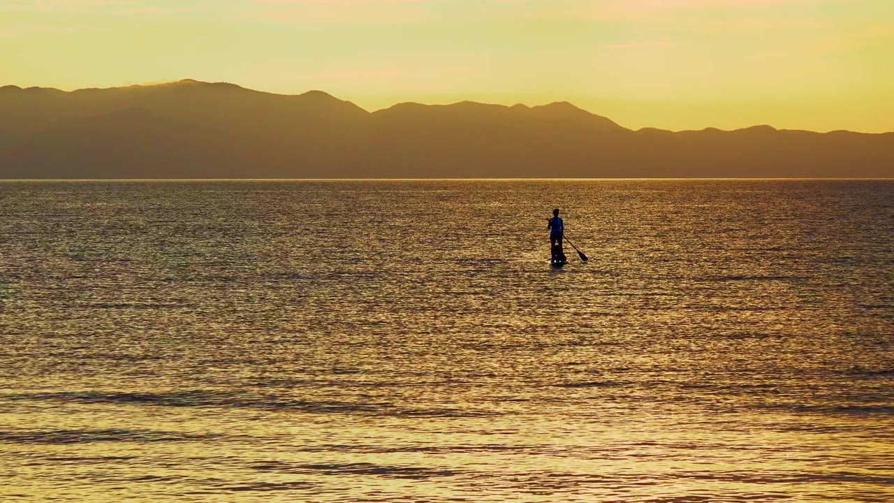 A silhouetted paddleboarder stands small in frame on golden ocean water in El Sargento La Ventana, Mexico, with dark mountains forming the distant horizon.