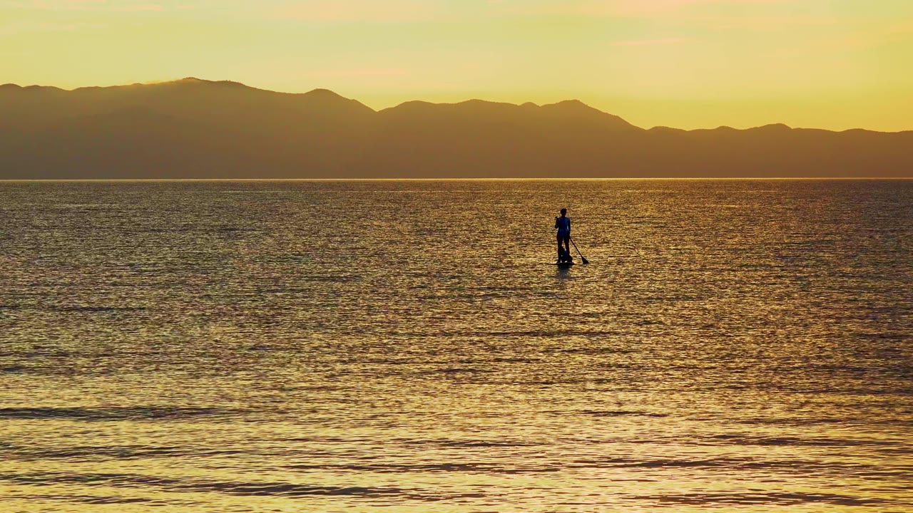 The paddleboarder appears slightly right of center on the golden water in El Sargento La Ventana, Mexico, with textured ripples leading back to the mountain horizon.