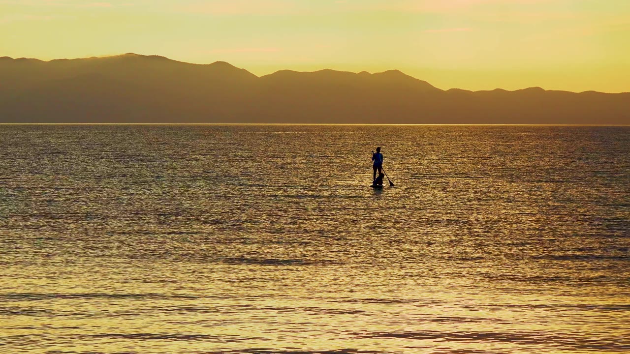 At 0:00, a distant silhouetted paddleboarder stands on reflective golden water in El Sargento La Ventana, Mexico, with dark mountains under a warm sky.