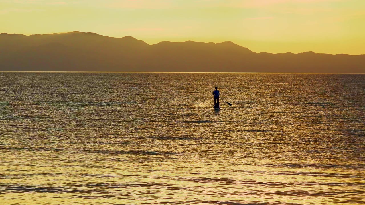Lone paddleboarder centered on shimmering water below a dark mountain horizon.