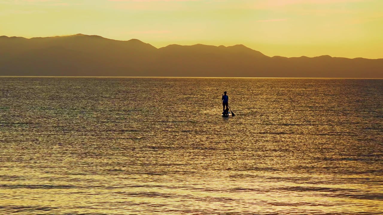 At 0:02, the lone paddleboarder appears slightly right of center on calm golden water in El Sargento La Ventana, Mexico, backed by a continuous dark mountain line.