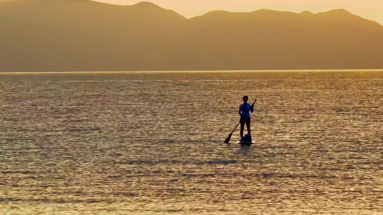 A paddleboarder stands left of center on calm golden water in La Ventana, Mexico, with dark mountains lining the sunset horizon.