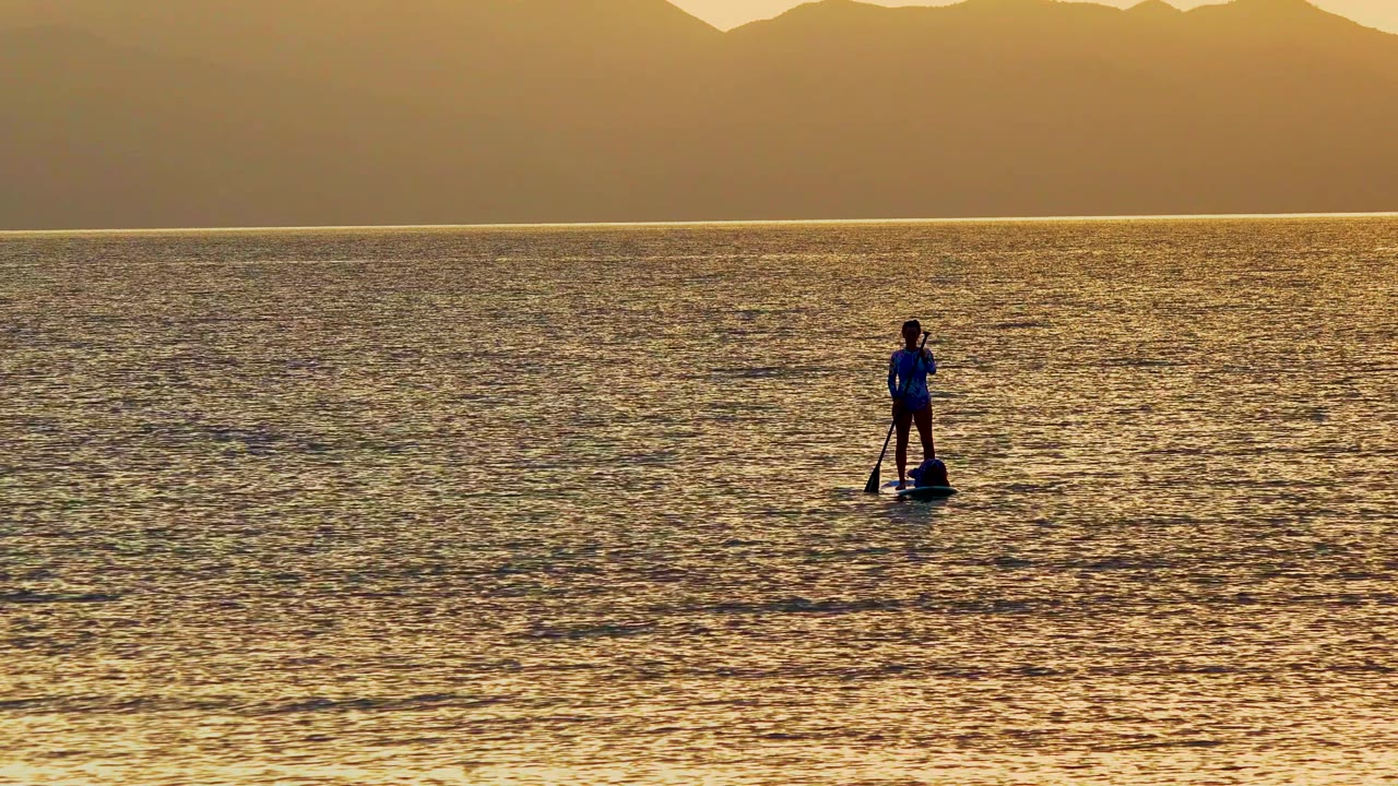 A paddleboarder stands near the right side of the frame on calm water in La Ventana, Mexico, with the paddle vertical and sunset mountains in the distance.