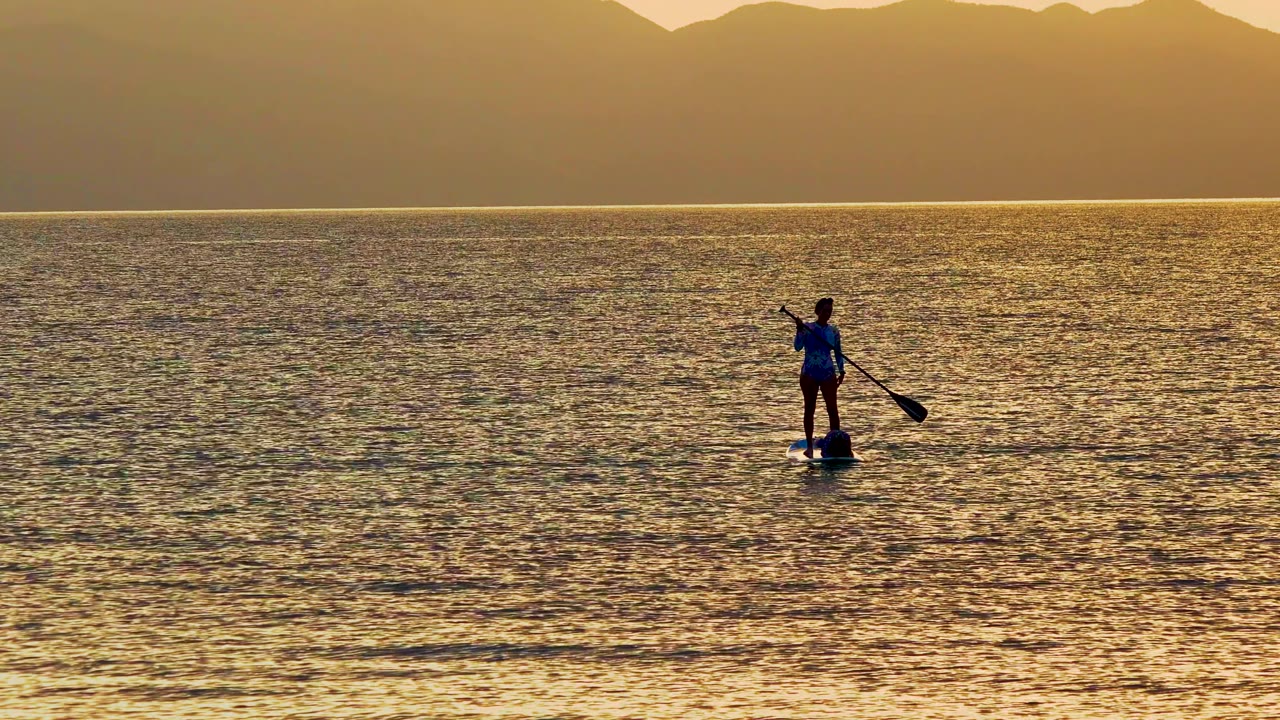 A lone paddleboarder stands near the left side of the frame on calm golden water in La Ventana, Mexico, with a dark mountain ridge along the horizon.