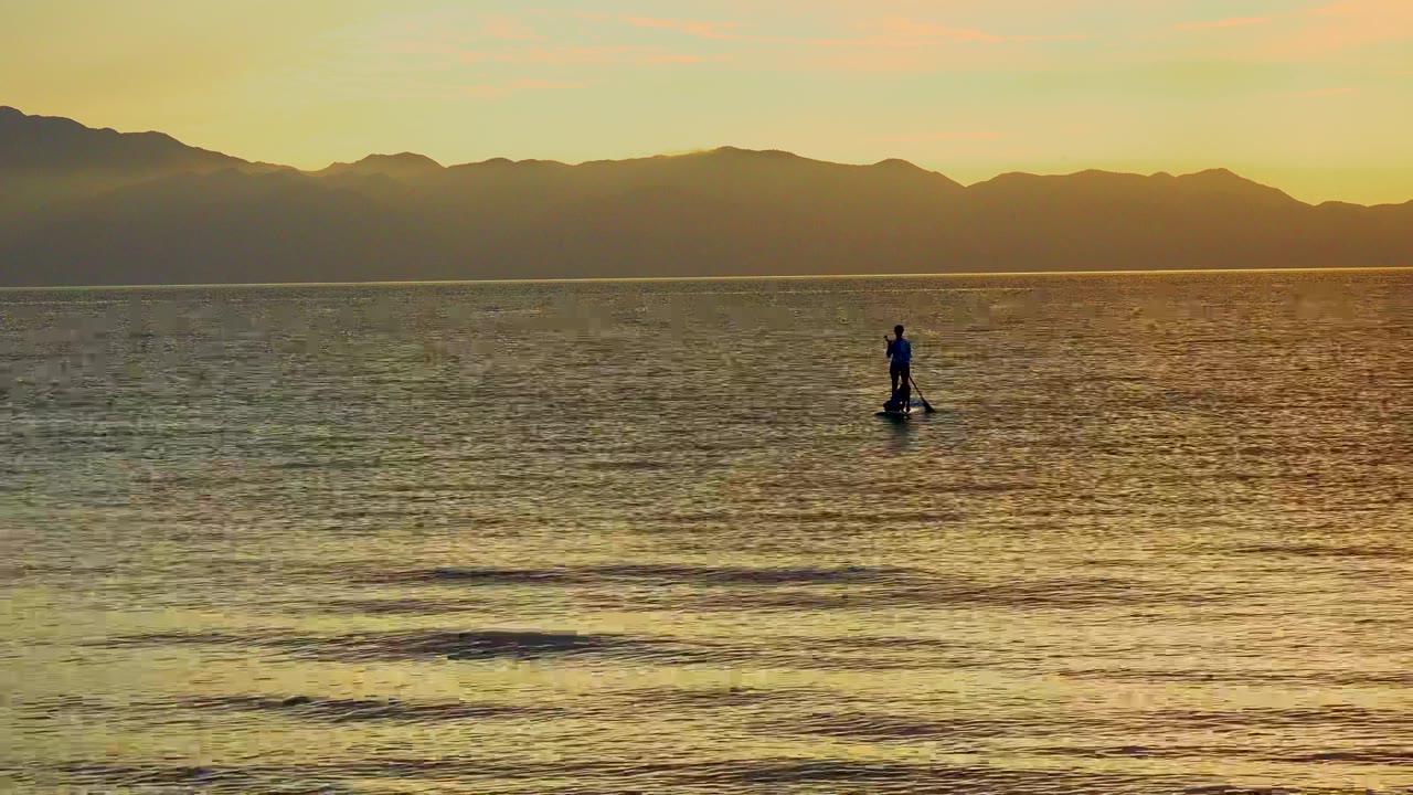 A distant paddleboarder stands on calm golden water in El Sargento La Ventana, Mexico, with dark mountains stretching across the horizon.