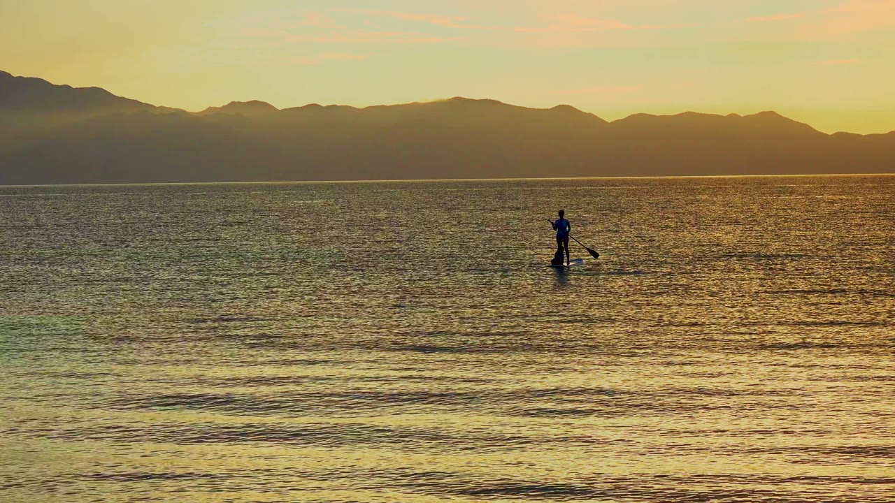 Distant paddleboarder crossing rippled sunset water.