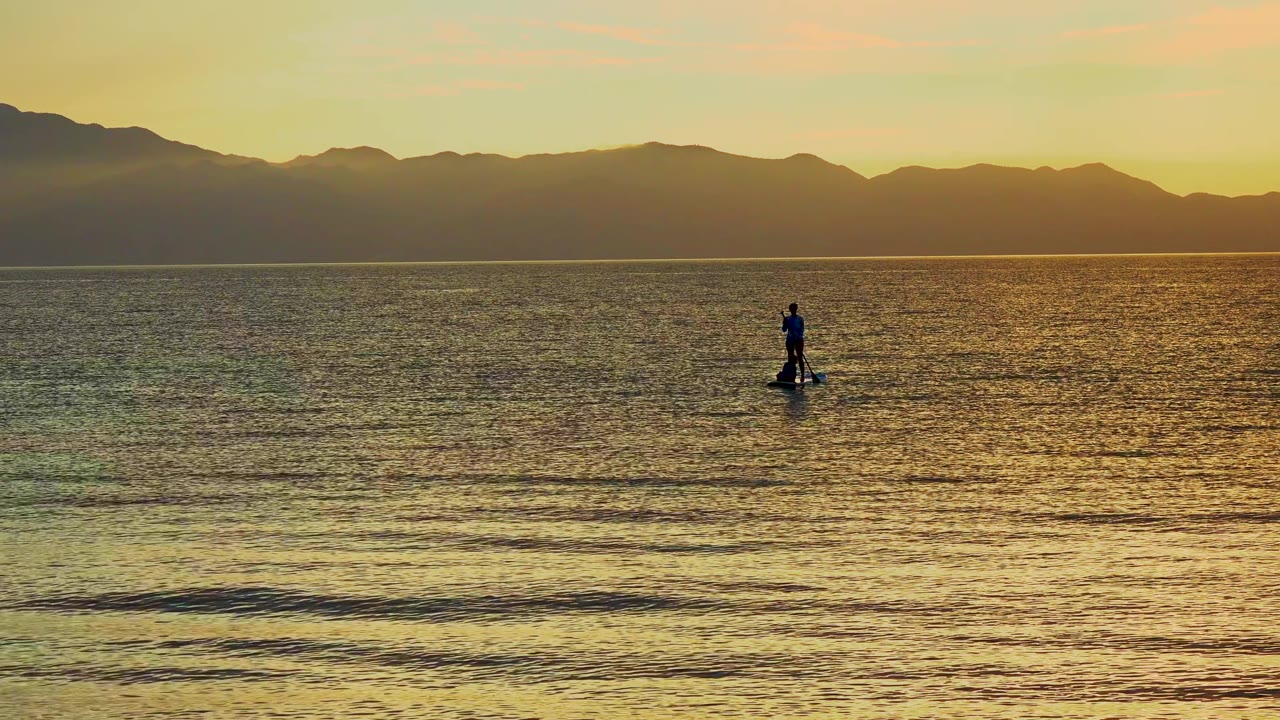 A small paddleboarder silhouette stands on calm ocean water in El Sargento La Ventana, Mexico, with amber reflections and a dark ridgeline beyond.