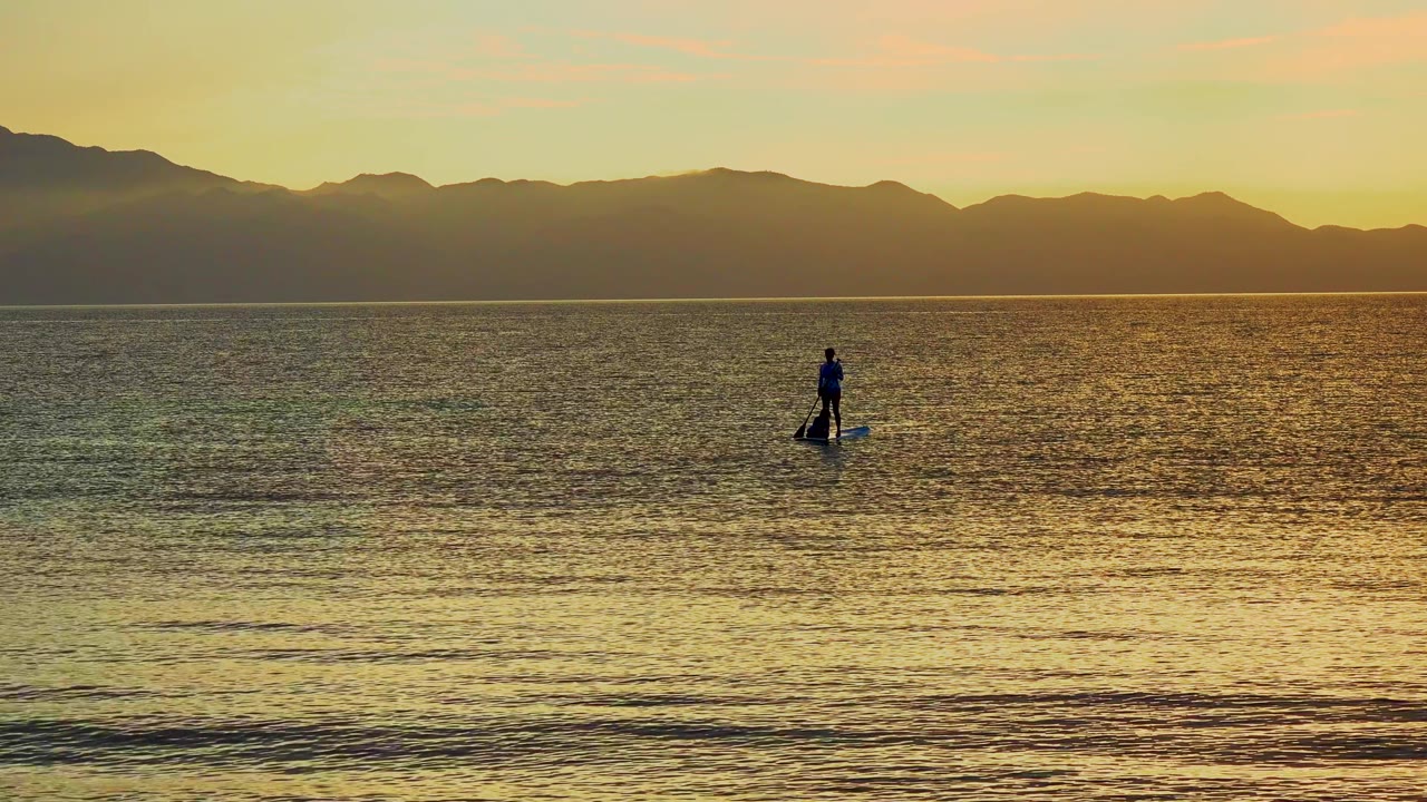 A distant paddleboarder stands on calm water in La Ventana, Mexico, with golden reflections, low foreground ripples, and dark mountains across the horizon.