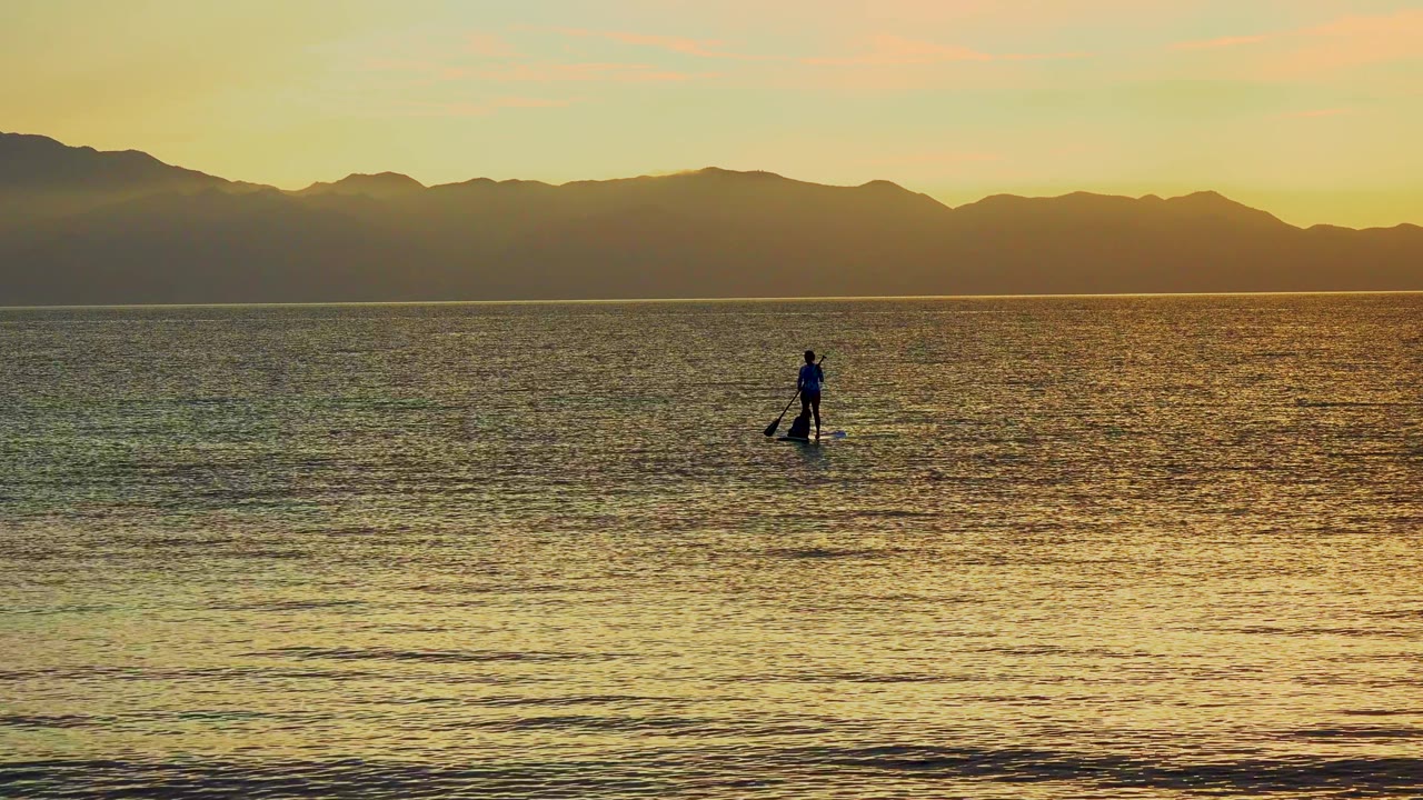 Paddleboarder on calm ocean as warm light reflects below mountains.