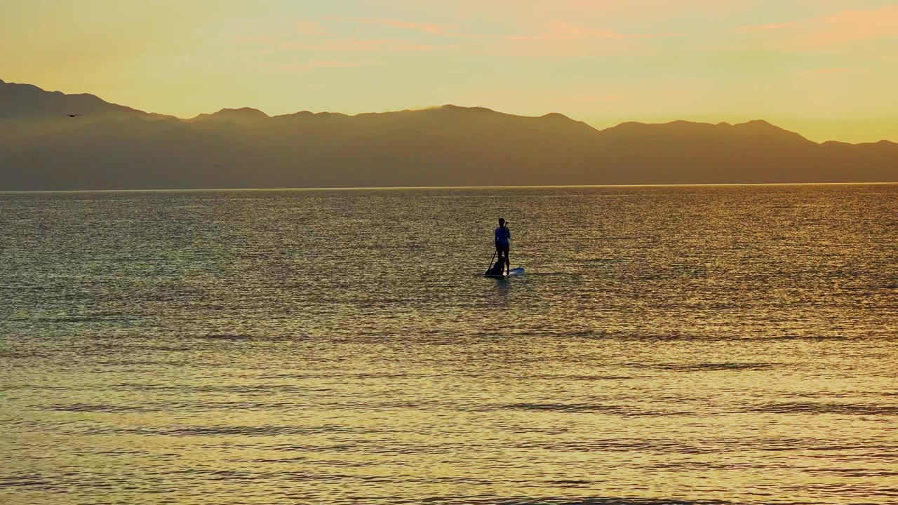 A small figure on a paddleboard is set farther right in the wide La Ventana frame, surrounded by gold and green water tones beneath the silhouetted mountains.