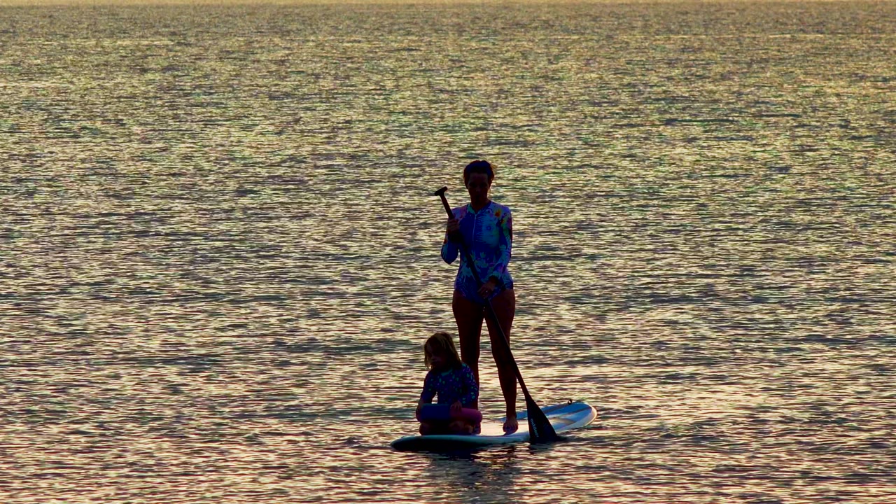 At 0:00 in La Ventana, Mexico, a silhouetted paddleboarder stands near the left side of the frame while a child sits on the board over gold rippled water.