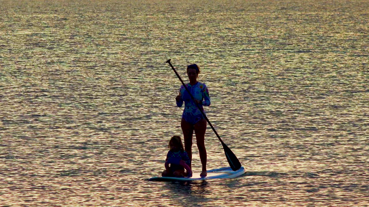 At 0:01 in La Ventana, Mexico, the standing rider holds the paddle more upright as the seated child remains near the front half of the board on calm water.