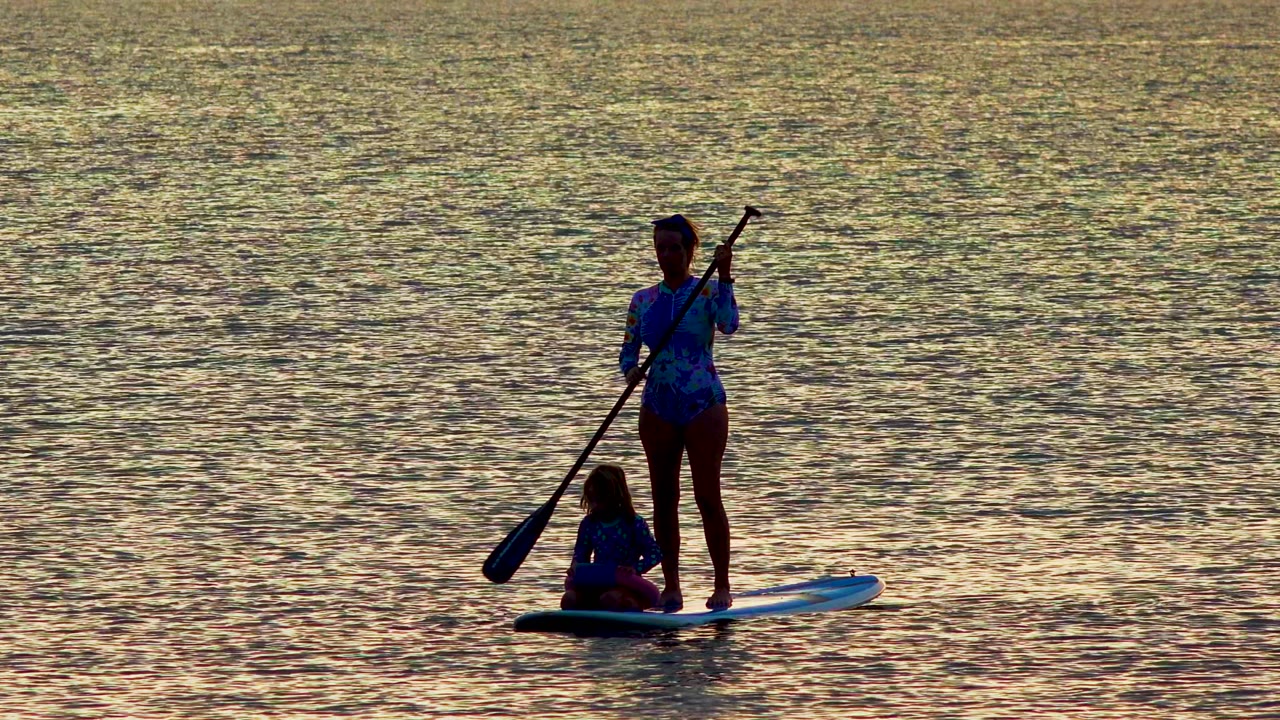At 0:02 in La Ventana, Mexico, the paddleboard sits toward the right of the frame with the standing rider holding the paddle across the body and the child seated low on the board.