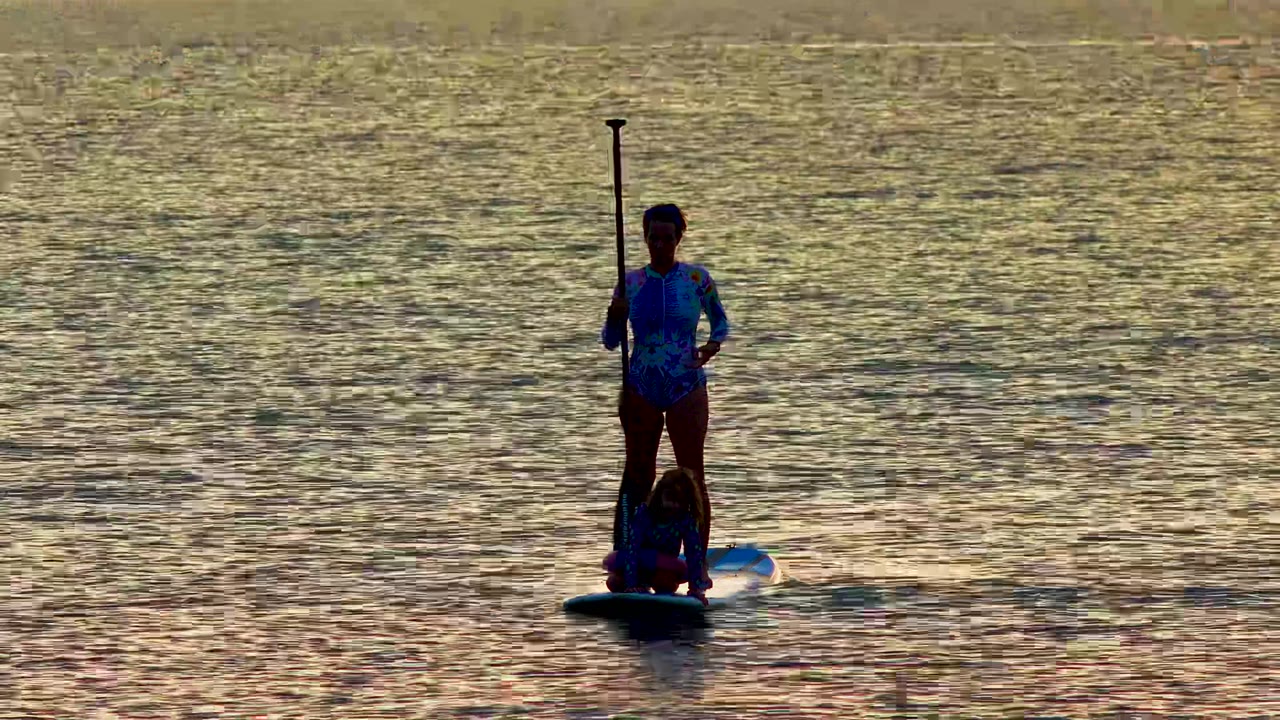Two people on a stand-up paddleboard in La Ventana, Mexico, with the standing person holding the paddle upright against reflective sunset water.