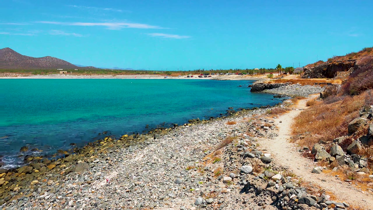 Wide view of the El Sargento La Ventana shoreline with turquoise water on the left, a pebbled beach below, a sandy path on the right, distant hills, and no visible people.