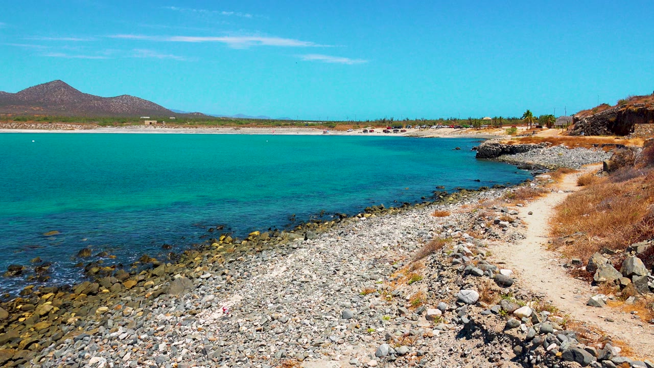 A bright El Sargento La Ventana shoreline view shows more open turquoise water, a pale rocky beach, a narrow path along dry vegetation, distant hills, and no visible people.