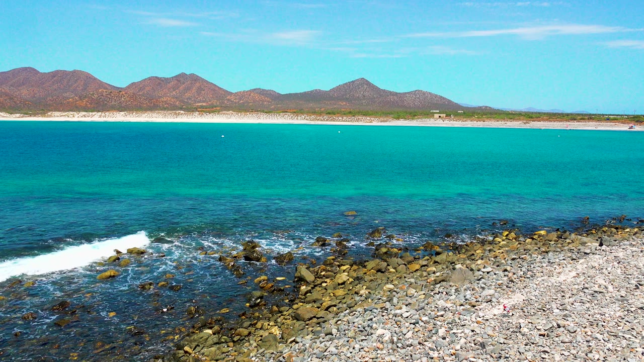 Rocky shoreline with white surf, turquoise ocean, beach, mountains, and clear sky.