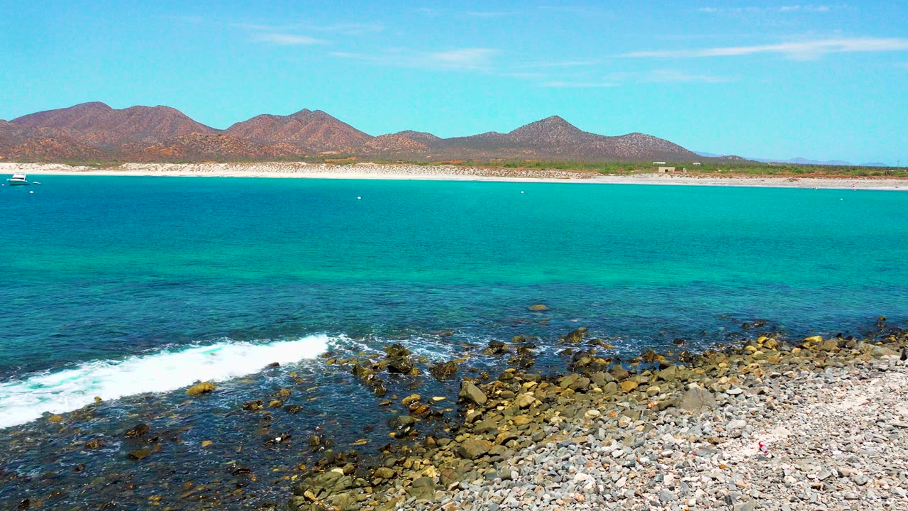At El Sargento La Ventana, a band of foam pushes farther across the rocky foreground below turquoise water, beach, distant mountains, clear sky, and no visible people.