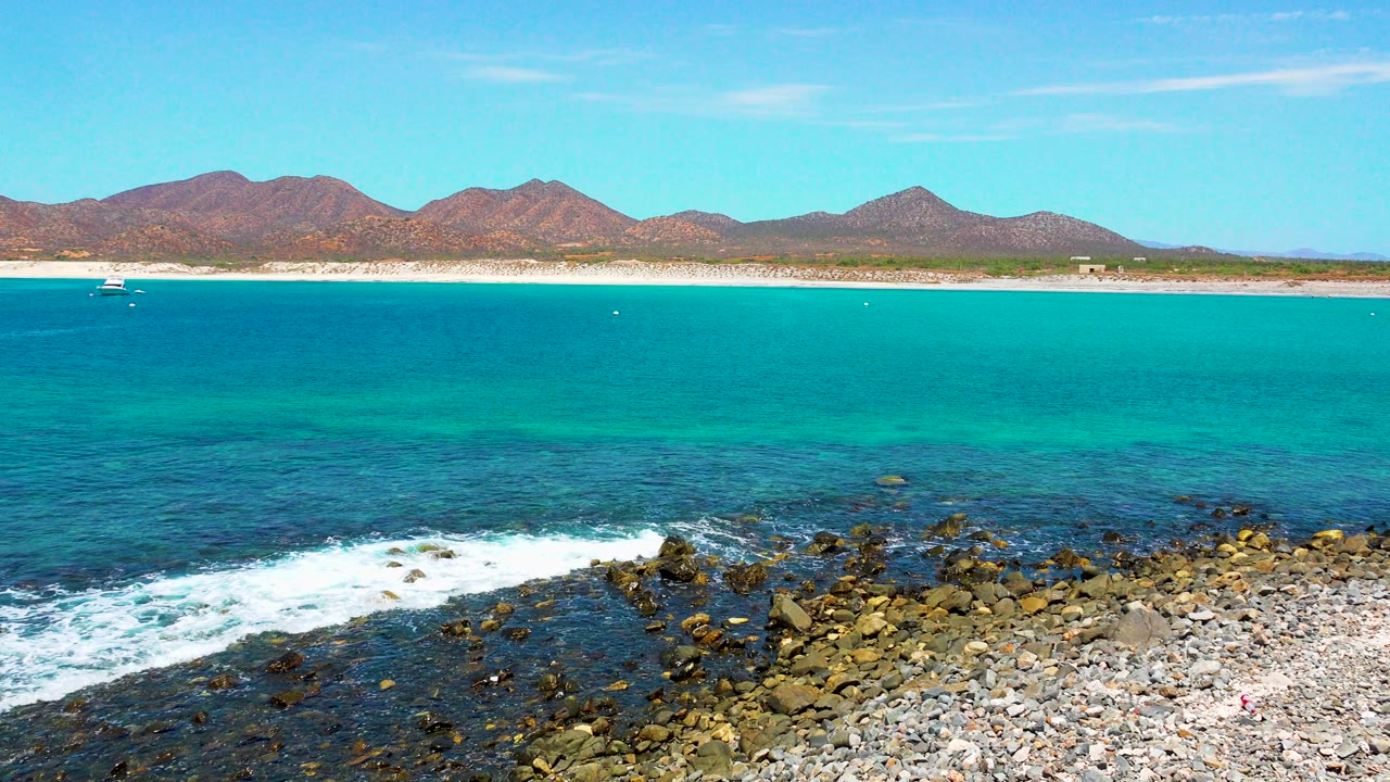 At El Sargento La Ventana, white water breaks along the left side of the rocky shore while the blue-green ocean, narrow beach, mountain horizon, and no visible people remain in view.