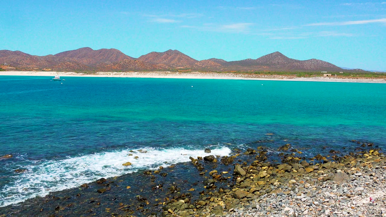 At El Sargento La Ventana, a small boat sits offshore beyond turquoise water as white surf washes the rocks, with beach, mountains, clear sky, and no visible people in the frame.
