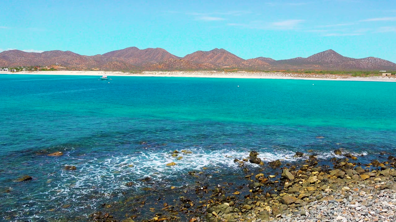 Opening frame of El Sargento La Ventana, Mexico with turquoise water, white foam on foreground rocks, a sandy beach, distant brown mountains, a small offshore boat, and no visible people.
