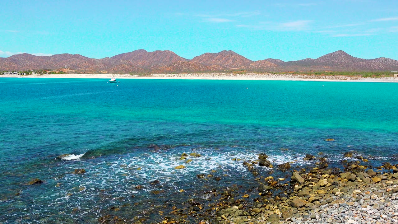 At one second in El Sargento La Ventana, Mexico, the white surf spreads wider over the dark shoreline rocks while the turquoise bay, pale beach, distant mountains, small boat, and no visible people remain in view.