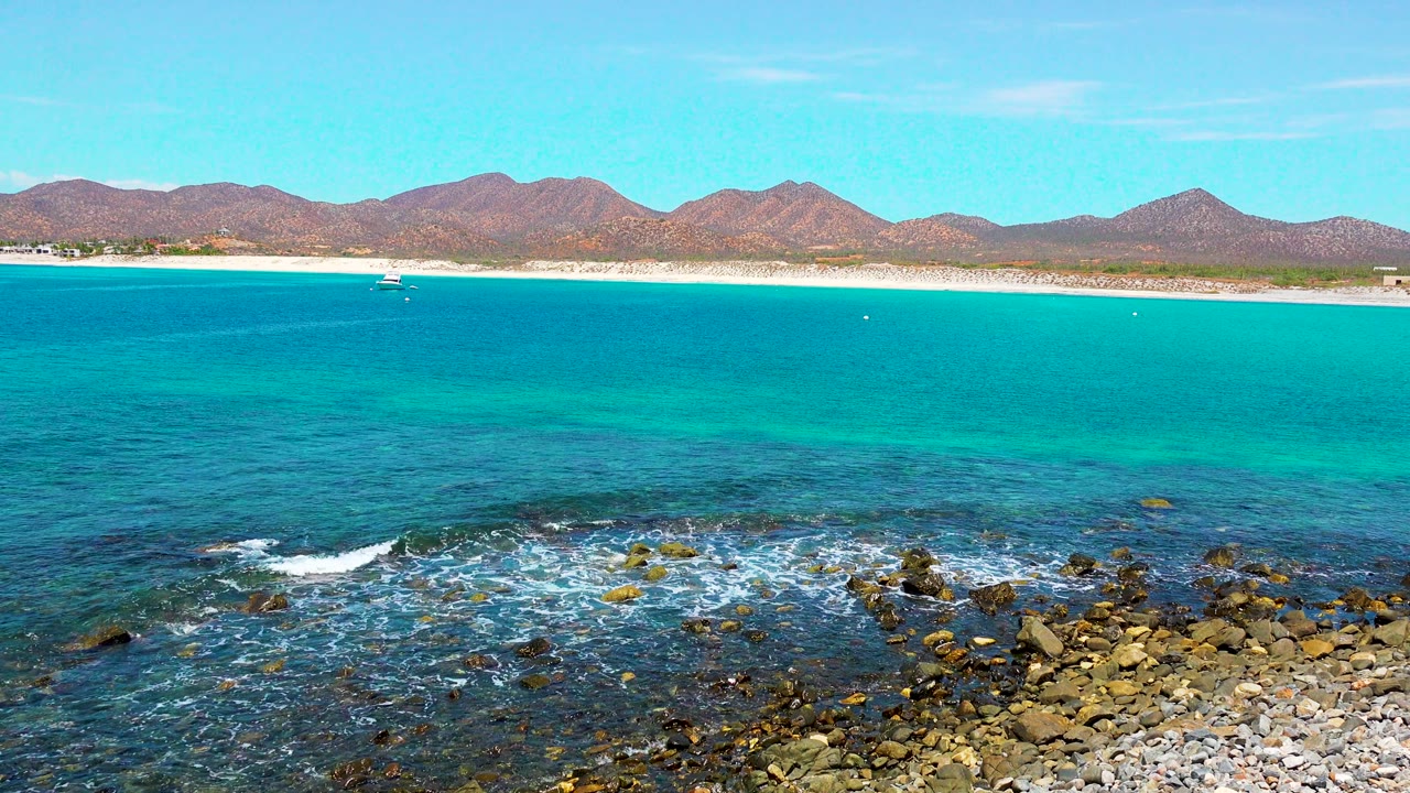 At two seconds, El Sargento La Ventana, Mexico remains framed with rocky foreground surf, clear turquoise water, the light beach strip, distant mountains, a small boat near shore, and no visible people.