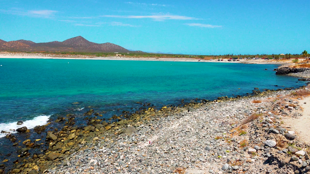 Opening frame of El Sargento La Ventana, Mexico, showing turquoise ocean, a pebbled rocky foreground, a pale beach strip, distant mountains, bright blue sky, and no visible people.