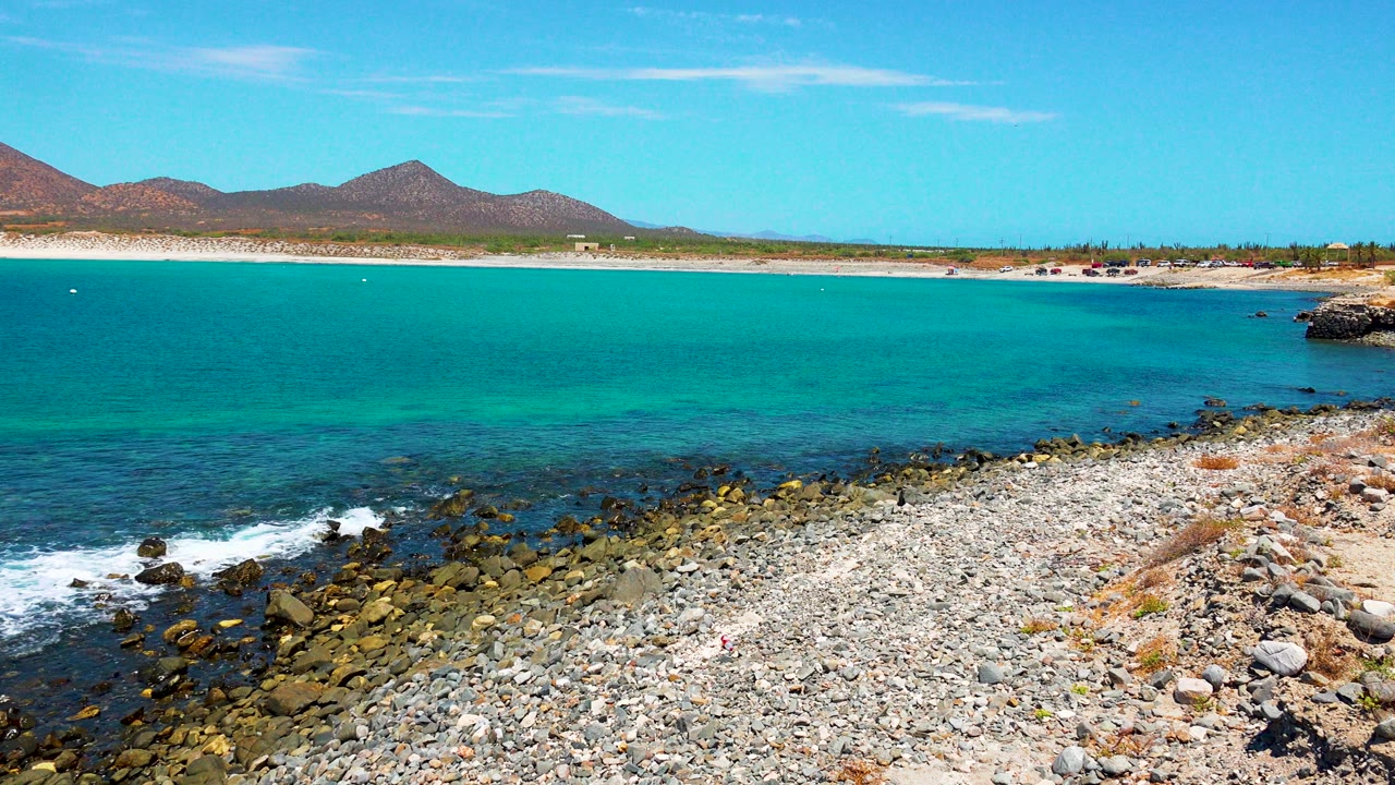 At 0:01 in El Sargento La Ventana, Mexico, a small patch of white foam pushes toward dark shoreline rocks below turquoise water, with beach and mountains in the distance and no visible people.