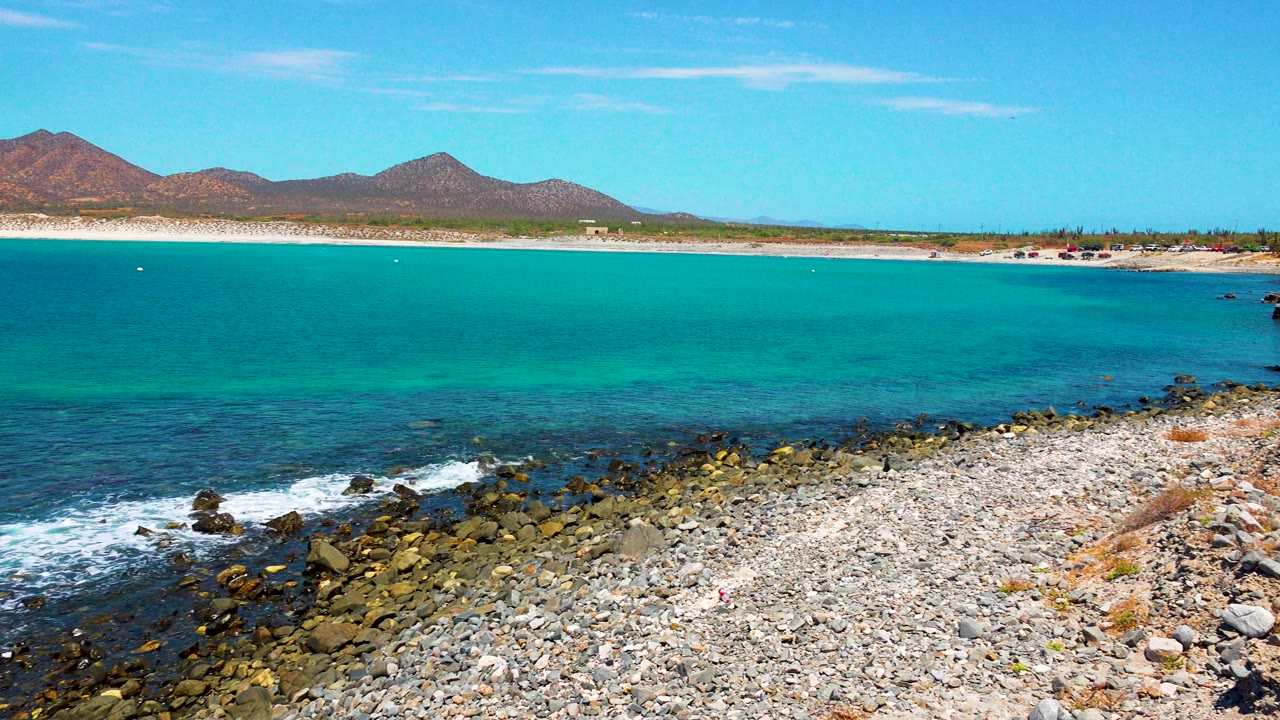 At 0:02 in El Sargento La Ventana, Mexico, the rocky foreground fills more of the lower frame as blue-green water stretches to a pale beach and low mountains, with no visible people.
