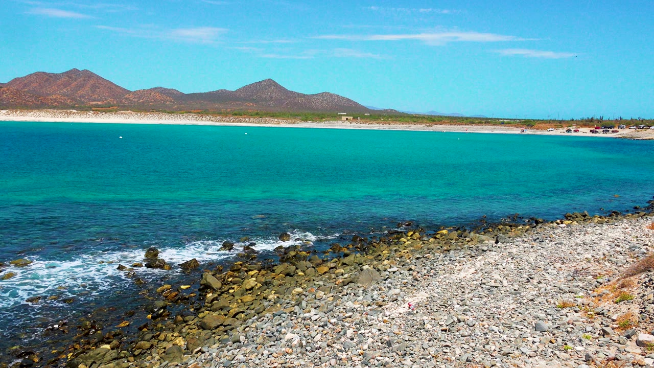 At 0:03 in El Sargento La Ventana, Mexico, white foam spreads along the lower left rocks while the turquoise bay, sandy shoreline, mountain ridge, and bright sky remain visible, with no visible people.