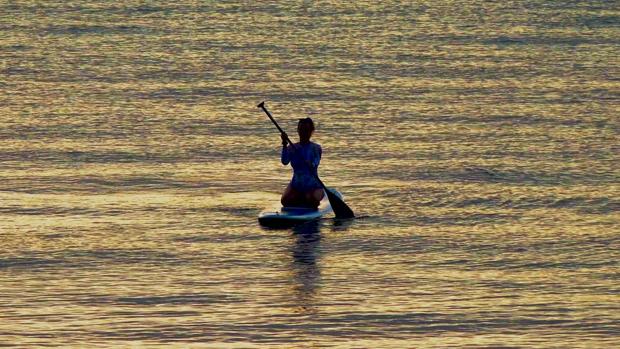 A silhouetted person kneels on a paddleboard in El Sargento, Mexico, with the paddle nearly vertical and a dark reflection beneath the board.