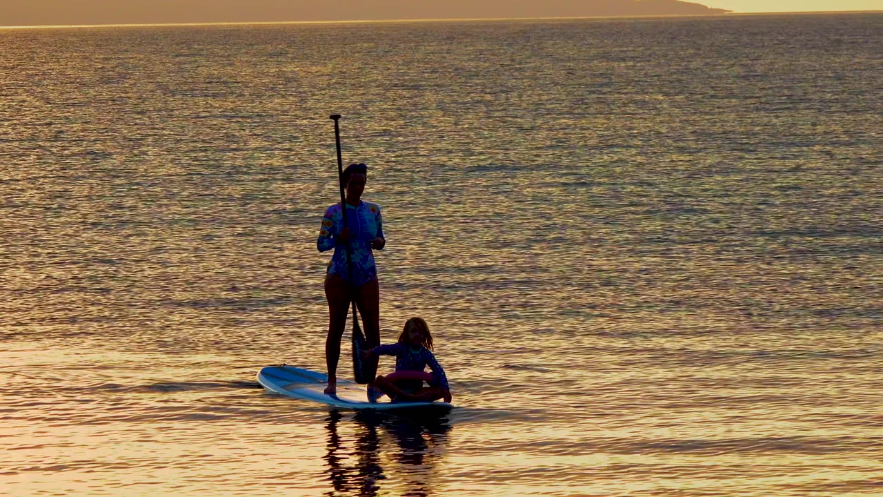 At 0:00 in El Sargento, Mexico, a woman stands upright on a blue paddleboard while a child stays low beside her on golden sunset water.