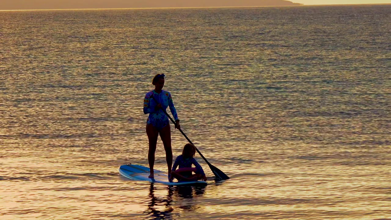 At 0:02 in El Sargento, Mexico, the silhouetted woman places the paddle toward the right side of the board while the child stays close to the deck.