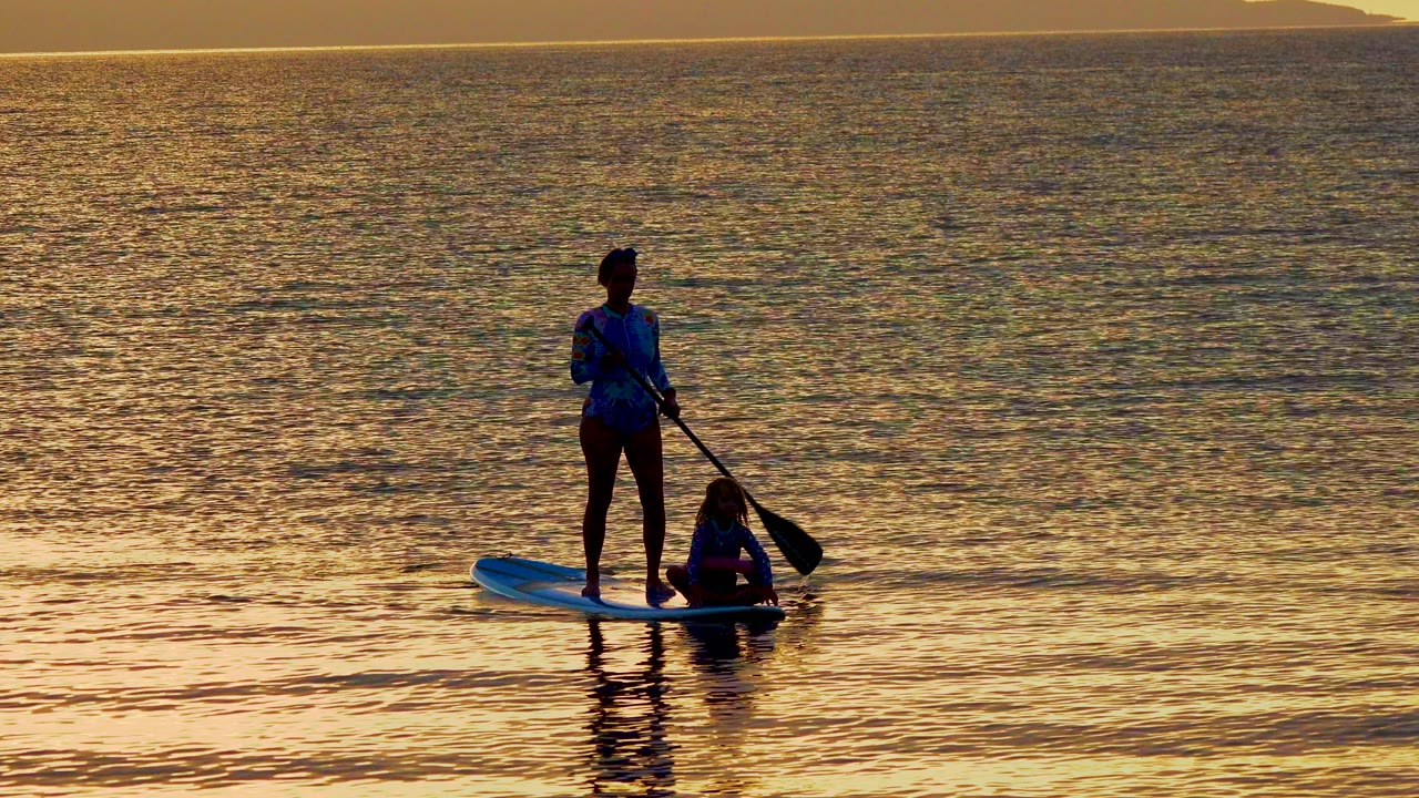 Standing paddleboarder beside a seated person on calm sunset water.