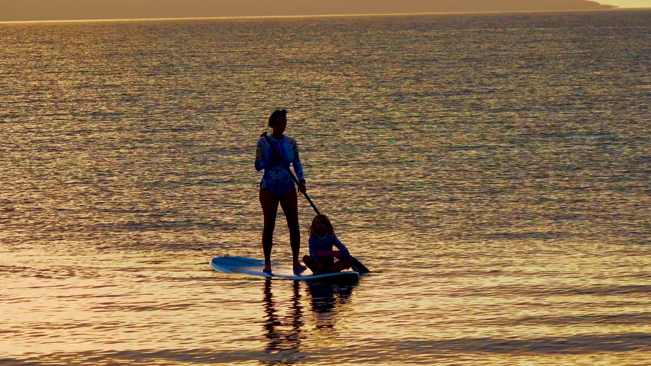 At 0:02 in El Sargento, Mexico, the standing paddleboarder faces forward while the second person sits lower on the board, both silhouetted against a broad band of golden ocean.